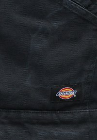 Μαύρο υφασμάτινο υλικό με μια μικρή ορθογώνια ετικέτα λογότυπου Dickies ραμμένη κοντά σε ορατές ραφές.