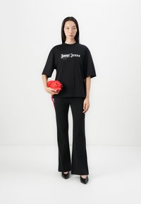 T-shirt noir oversize avec du texte scintillant "Joon Jeans", associé à un pantalon évasé noir. Le mannequin tient une pochette en satin rouge.