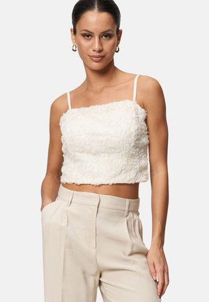 White cropped top with textured floral detailing, thin adjustable straps; paired with beige high-waisted trousers for a contrasting look.