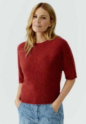 Vrouw met lang blond haar, gekleed in een rood, pluizig, kortmouwig truitje en blauwe spijkerbroek met paisleypatroon, staand met handen in de zakken.