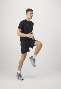 Camiseta deportiva negra y pantalones cortos, zapatillas de correr grises. Modelo en movimiento, levantando una rodilla con un fondo neutro.