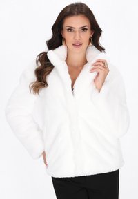 Kvinde iført en hvid faux fur frakke, med langt bølget hår, guldøreringe og guld ringe, der poserer mod en ensfarvet baggrund.