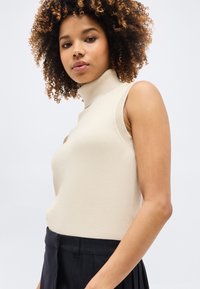 Γυναίκα με κατσαρά μαλλιά που φοράει ένα μπεζ αμάνικο τοπ με λαιμόκοψη και σκούρο παντελόνι ψηλόμεσο, ποζάροντας μπροστά σε ένα απλό ανοιχτό φόντο.