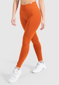 Leggings arancioni con un alto elastico in vita, realizzati in un materiale liscio e stretch. Indossati con scarpe da ginnastica bianche. Design semplice, senza fantasie.