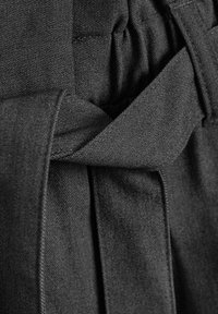 Ceinture en tissu noir avec un détail de nœud, mettant en avant un tissage texturé. Le matériau semble robuste et lisse, réfléchissant la lumière de manière subtile.