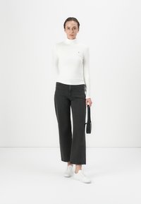 Weißes Langarm-Turtleneck, schwarze weite Jeans und weiße Sneaker. Das Model hält eine kleine schwarze Handtasche. Minimalistischer Hintergrund.