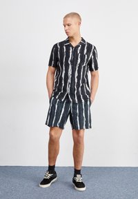 Camisa de botones con rayas verticales en blanco y negro, combinada con pantalones cortos a juego. Zapatillas negras con suelas blancas. Fondo gris.