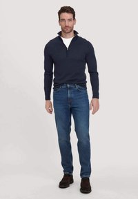 Marineblauwe half-zip trui gemaakt van een zachte stof, gecombineerd met een blauwe jeans en donkerbruine schoenen, die een casual, getailleerde silhouet laat zien.