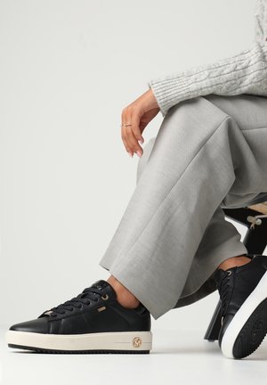 Henkilö istuu yhdellä jalalla ristissä, pukeutuneena vaaleanharmaisiin housuihin, harmaaseen palmikkoneulepaitaan ja mustiin tennareihin, joissa on valkoiset pohjat.