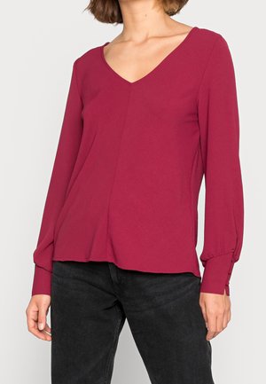 Vrouw draagt een dieproze V-hals blouse met lange mouwen en knoopmanschetten en zwarte broek, staand tegen een effen achtergrond.