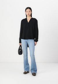 Top negro de manga larga con escote en V y detalle de botones, combinado con jeans acampanados de color azul claro y zapatos de estampado animal, sosteniendo un pequeño bolso negro.