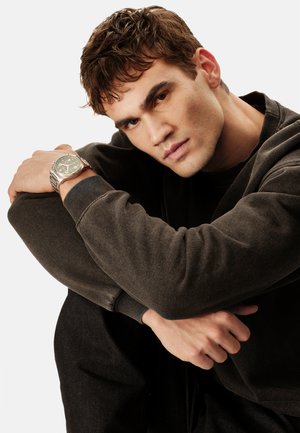 Jeune homme aux cheveux bruns portant un sweat-shirt foncé, les bras reposant sur les genoux pliés, portant une montre-bracelet argentée au cadran sombre.