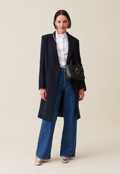 Cappotto navy, blusa bianca con volant, jeans in denim blu a gamba larga e scarpe metalliche. Una piccola borsa nera con un dettaglio ad anello dorato.