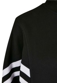 Svart sweatshirt med hög krage. Har texturerad tyg och tre horisontella vita ränder på ärmarna. Sömmar är synliga.