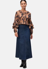 Brown blouse with black abstract pattern, ruffled cuffs, and tie neck; dark blue denim maxi skirt with side seams; black ankle boots.
