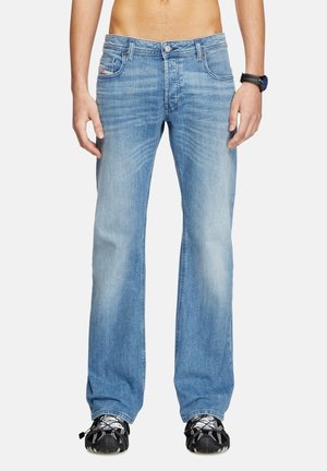 Homme portant un jean droit bleu clair, des baskets noires et une montre noire au poignet, montré du milieu du torse jusqu’aux pieds sur fond blanc.