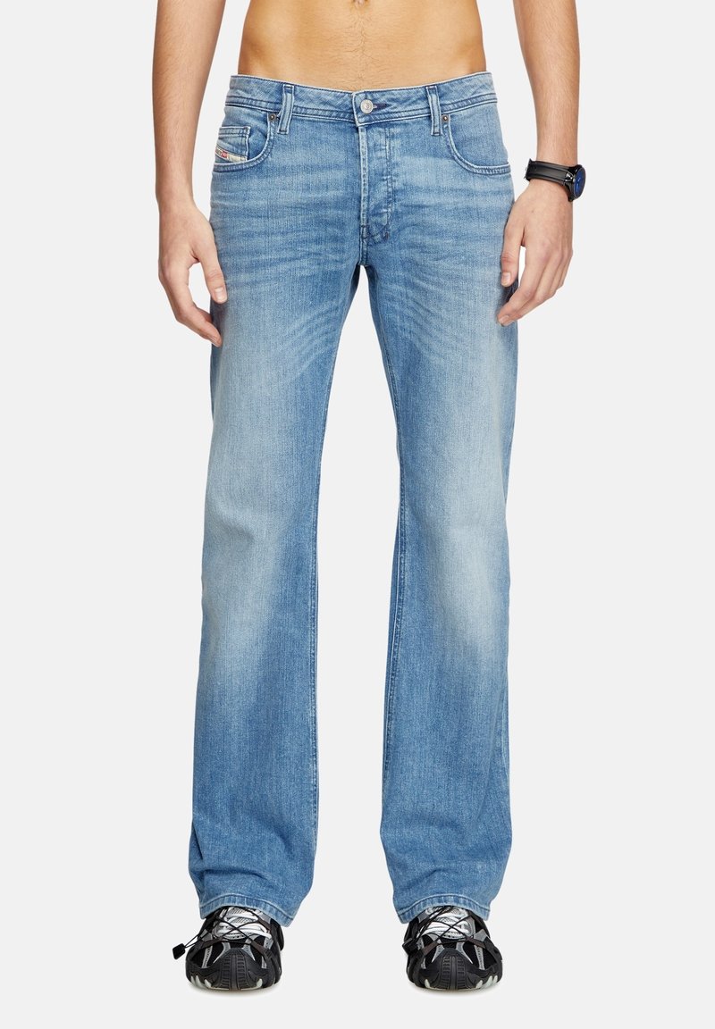Homme portant un jean droit bleu clair, des baskets noires et une montre noire au poignet, montré du milieu du torse jusqu’aux pieds sur fond blanc.