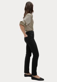 Zwarte jeans met een slim fit, gecombineerd met een beige en zwart gestreept blouse. De vrouw staat zijwaarts, met haar handen in de zakken, en draagt platte schoenen.
