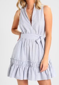 Vestido sin mangas a rayas en azul claro y blanco, con un profundo escote en V, una cintura ceñida con cinturón y un dobladillo con volantes y detalles fruncidos.