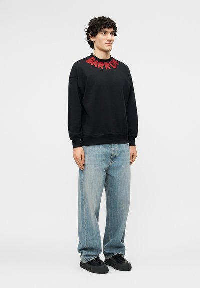 Νεαρός με σγουρά μαλλιά, που φοράει μαύρο φούτερ με κόκκινο κείμενο στο γιακά, ανοιχτό μπλε φαρδιά τζιν και μαύρα αθλητικά παπούτσια, στέκεται όρθιος.