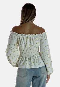 Weiße schulterfreie Bluse mit gelbem Blumenmuster, gerüschtem Taillenbereich, langen Puffärmeln und rüschigem Ausschnitt. Getragen mit blauen Jeans.
