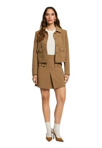 Chaqueta de color beige con botones dobles y bolsillos frontales, combinada con una falda a juego. El conjunto incluye calcetines beige y tacones altos marrones.