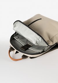 Zaino con un'esterno beige, interno grigio, dettagli in pelle e un comparto con zip. Presenta spallacci imbottiti e tasche interne.