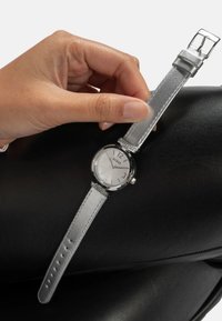 Hõbedane randme kell, millel on ümmargune, selge kristalliga kellapea, hõbedane rihm ja dekoratiivsed aktsendid rihma lähedal. Nähtav on käsi, mis reguleerib rihma.