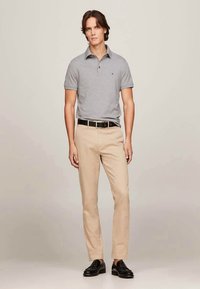 Jovem de pé a usar uma camisa polo cinza, calças bege, cinto preto e sapatos de pele pretos sobre um fundo simples.