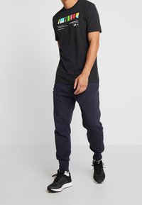 Svart t-shirt med färggrafisk tryck på framsidan, tillsammans med marinblå träningsbyxor. Modell bär svarta löparskor och står på en ljus bakgrund.