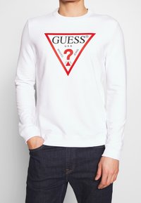 Sweatshirt bianco a maniche lunghe con un design del logo triangolare rosso con la scritta "GUESS" e un punto interrogativo al centro.