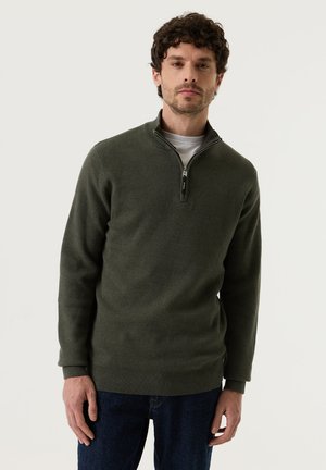 Mann mit kurzen, lockigen Haaren trägt einen olivgrünen Quarter-Zip-Pullover über einem weißen T-Shirt und dunkelblauen Jeans und steht vor einem schlichten Hintergrund.