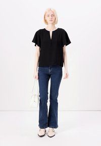 Black blouse with flutter sleeves, V-neckline, and buttons. Paired with flared jeans and light-colored shoes, holding a white bag.