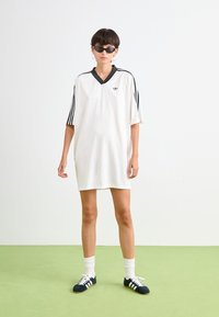 Balts sporta tērps ar melniem trīsrakstu akcentiem, ar V veida kakla izgriezumu, īsām piedurknēm un izšūtu logo. Valkāts ar melnām kurpēm un baltiem zeķēm.