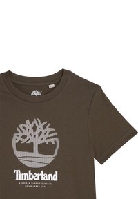 Hnědé tričko s krátkým rukávem vyrobené z bavlny, s bílou grafikou stromu a textem "Timberland". Jednoduchý kulatý výstřih, jemná textura.