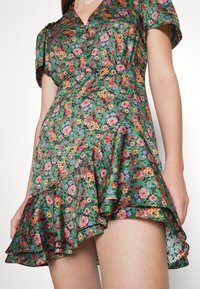 Robe courte verte à motif floral avec ourlet à volants et manches courtes portée par une personne debout sur un fond clair uni.