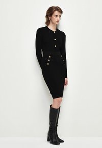 Schwarzes geripptes Kleid mit langen Ärmeln, Vorderknöpfen, figurbetontem Silhouette und goldenen Knopfdetails. Kombiniert mit knielangen schwarzen Stiefeln.