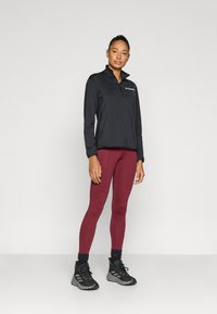 Fekete hosszú ujjú pulóver cipzáros gallérral, bordó leggingsszel, varrási részletekkel, és fekete sportcipővel párosítva.