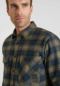 Geruite flanellen shirt in olijfgroen en marineblauw, met twee voorzakken, knoopsluiting en een gestructureerde stof met contrasterende patronen.