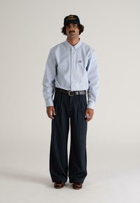 Man met een snor die een lichtblauw overhemd met knopen, donkerblauwe gestreepte wijde broek, een leren bruine riem, donkerbruine schoenen en een zwarte pet draagt.