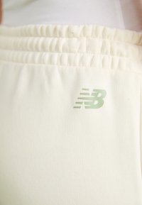 Krémové športové nohavice s textúrovanou látkou, s naťahovacím pásom a zeleným logom New Balance na ľavom stehne.