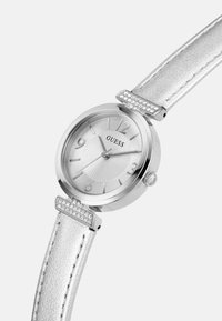 Hõbedane ümmargune kell tekstureeritud valge dials, kristallaktsentidega silmade ligidal ja metallist hõbedase nahkrihmaga.