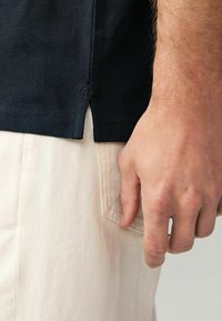Marineblå tekstureret skjorte med sidesplitter, parret med lyse beige bukser. Hånden placeret nær lommen fremhæver stofkontrast og farve.