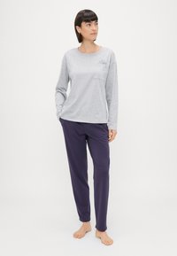 Dlhý rukáv sivý bavlnený top s vreckom a kvetinovou výšivkou; skombinovaný s voľnými tmavo purpurovými nohavicami vyrobenými z hladkého materiálu.