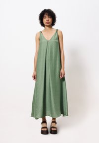 Grünes, ärmelloses Maxikleid mit V-Ausschnitt und gebundenen Trägern. Der strukturierte Stoff fällt locker und wird mit braunen Plateausandalen kombiniert.