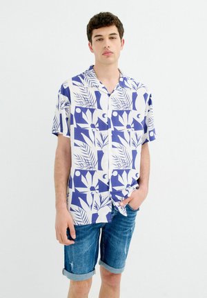 Joven con una camisa azul y blanca de manga corta con botones y pantalones cortos vaqueros remangados, de pie frente a un fondo liso.