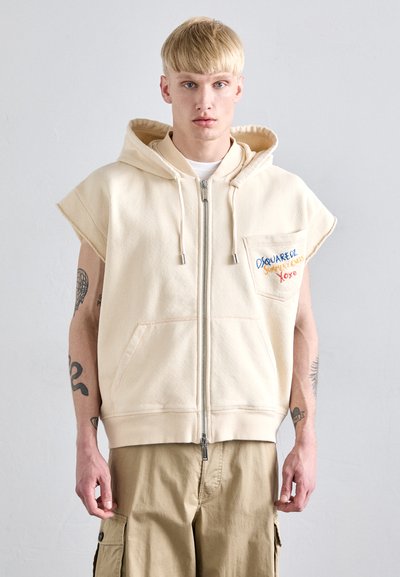Cremefarvet ærmeløs hoodie med frontlynlås, stor kængurulomme og broderet logo på brystet. Stoffet ser blødt og tekstureret ud.