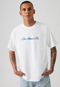 Weißes Baumwoll-T-Shirt mit blauem "Levi Strauss & Co." Textlogo. Kurze Ärmel und Rundhalsausschnitt. Getragen mit hellen Jeans.