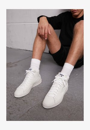 Beli visoki športni copati iz gladkega usnja z ravnimi vezalkami, teksturiranimi podplati in diskretnimi blagovnimi poudarki. Nosi se jih z belimi kratkimi nogavicami.