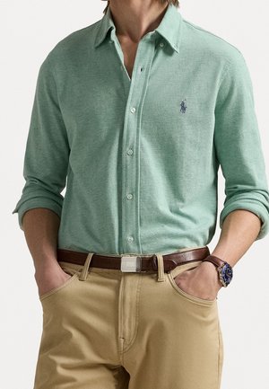 Man draagt lichtgroen overhemd met knopen en opgerolde mouwen, beige broek, bruine riem en polshorloge met blauwe wijzerplaat, handen in zijn zakken.